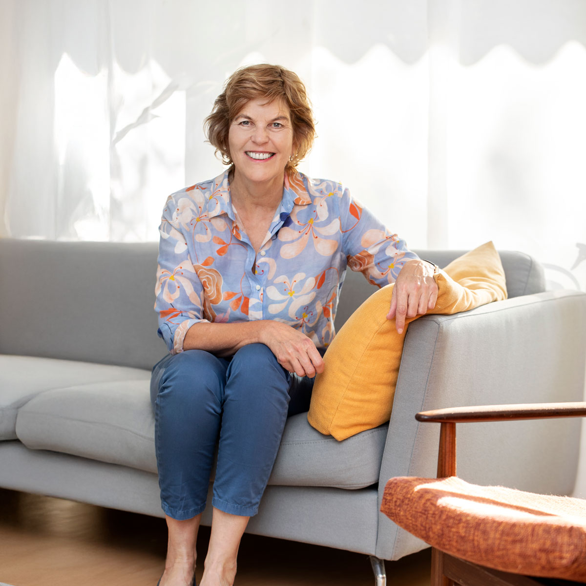 Frau entspannt sich auf grauem Sofa und l&auml;chelt in die Kamera.